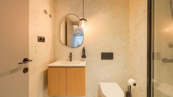 A modern bathroom with a marble wall, a wooden vanity, a round mirror, and a glass shower enclosure.