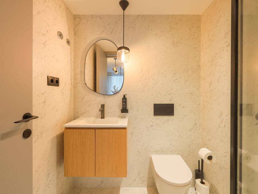 A modern bathroom with a marble wall, a wooden vanity, a round mirror, and a glass shower enclosure.