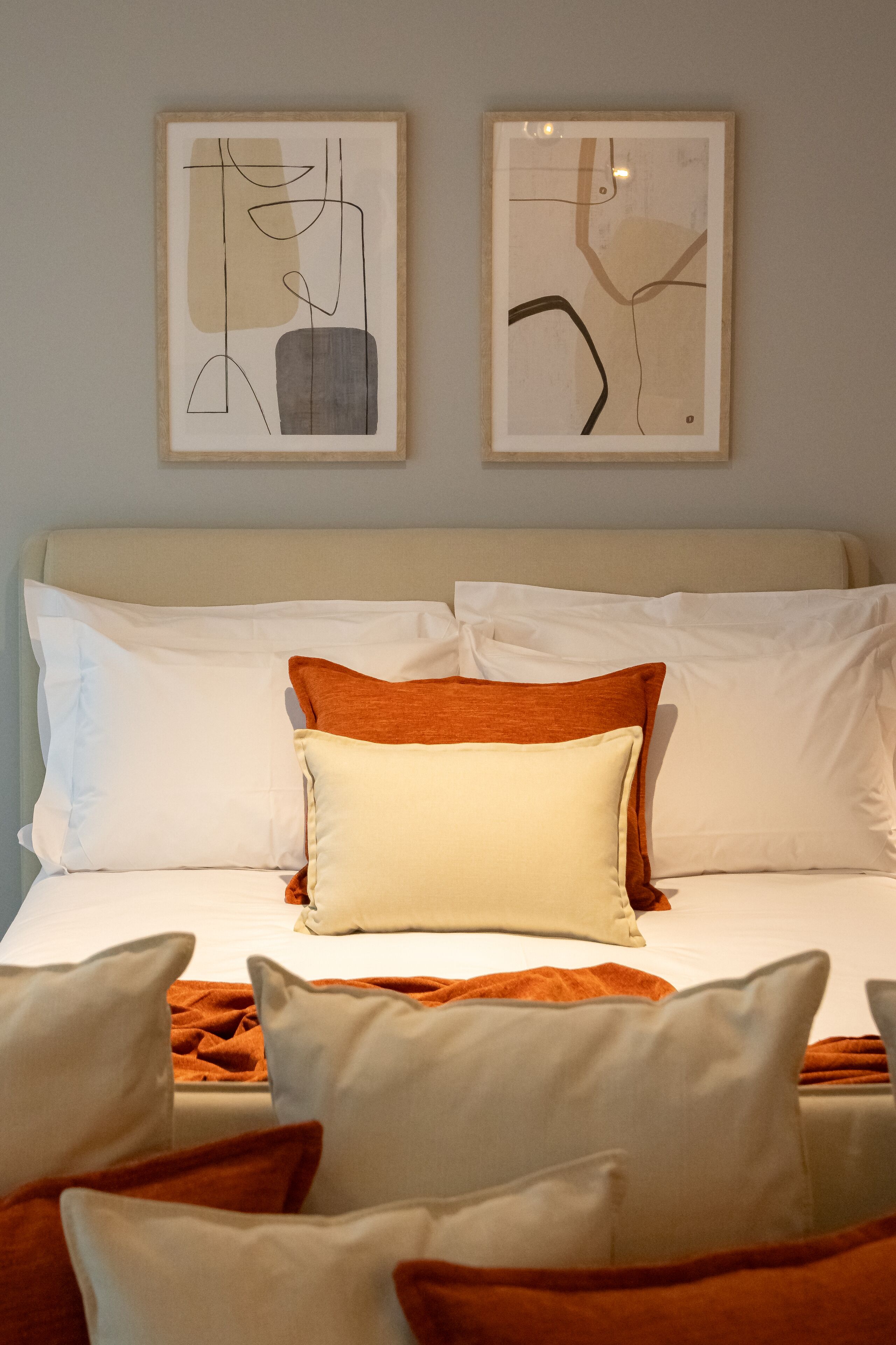 Ein ordentlich bezogenes Bett mit weißen und orangefarbenen Kissen, ein beigefarbener Kopfteil und ein abstraktes Kunstwerk an der Wand.