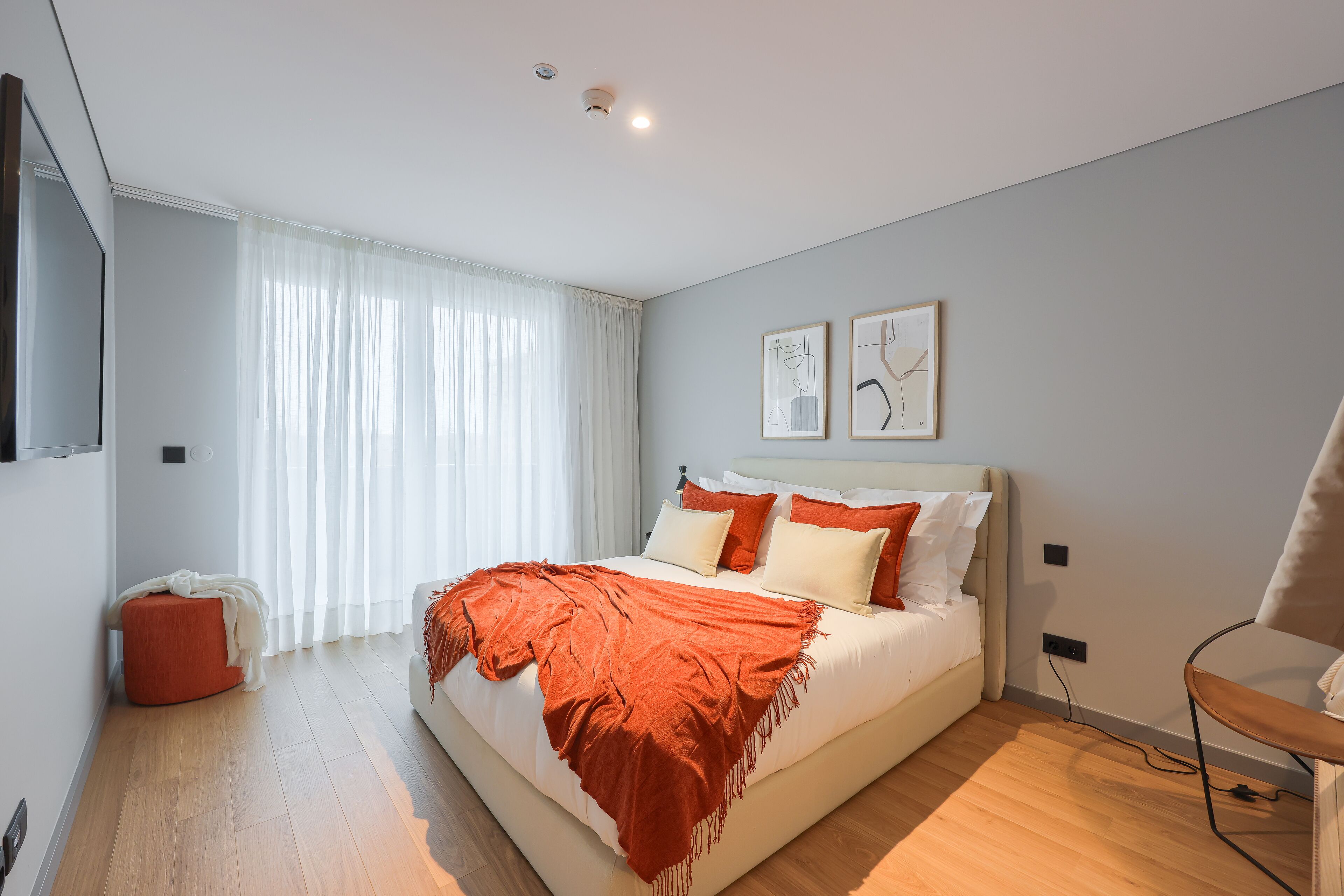 A modern bedroom with a large bed, a small ottoman, a chair, and artwork on the wall.