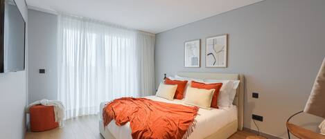 A modern bedroom with a large bed, a small ottoman, a chair, and artwork on the wall.