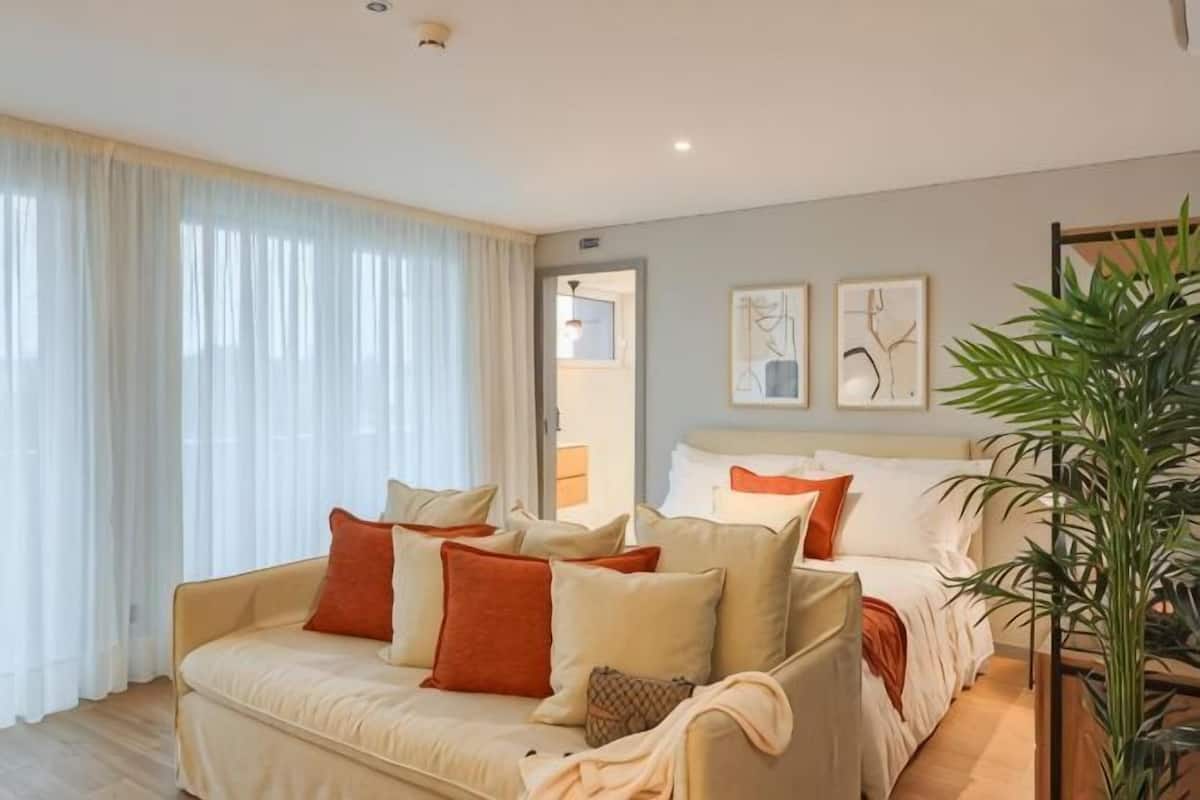 Una habitación de hotel moderna con un sofá color beige, una cama con ropa de cama blanca y naranja, un ventanal con cortinas blancas y una planta.