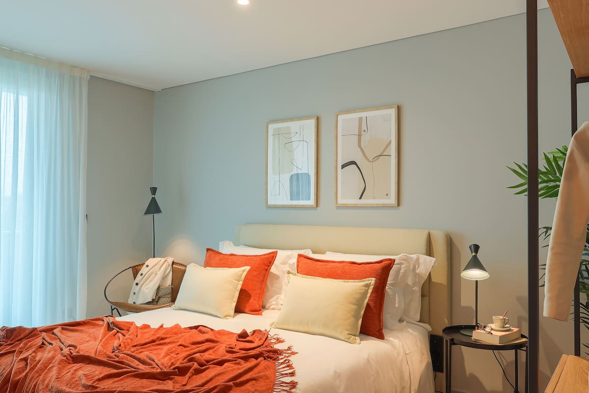 Una cama bien hecha con ropa de cama naranja y blanca, una mesita de noche con una lámpara y dos obras de arte enmarcadas en la pared.