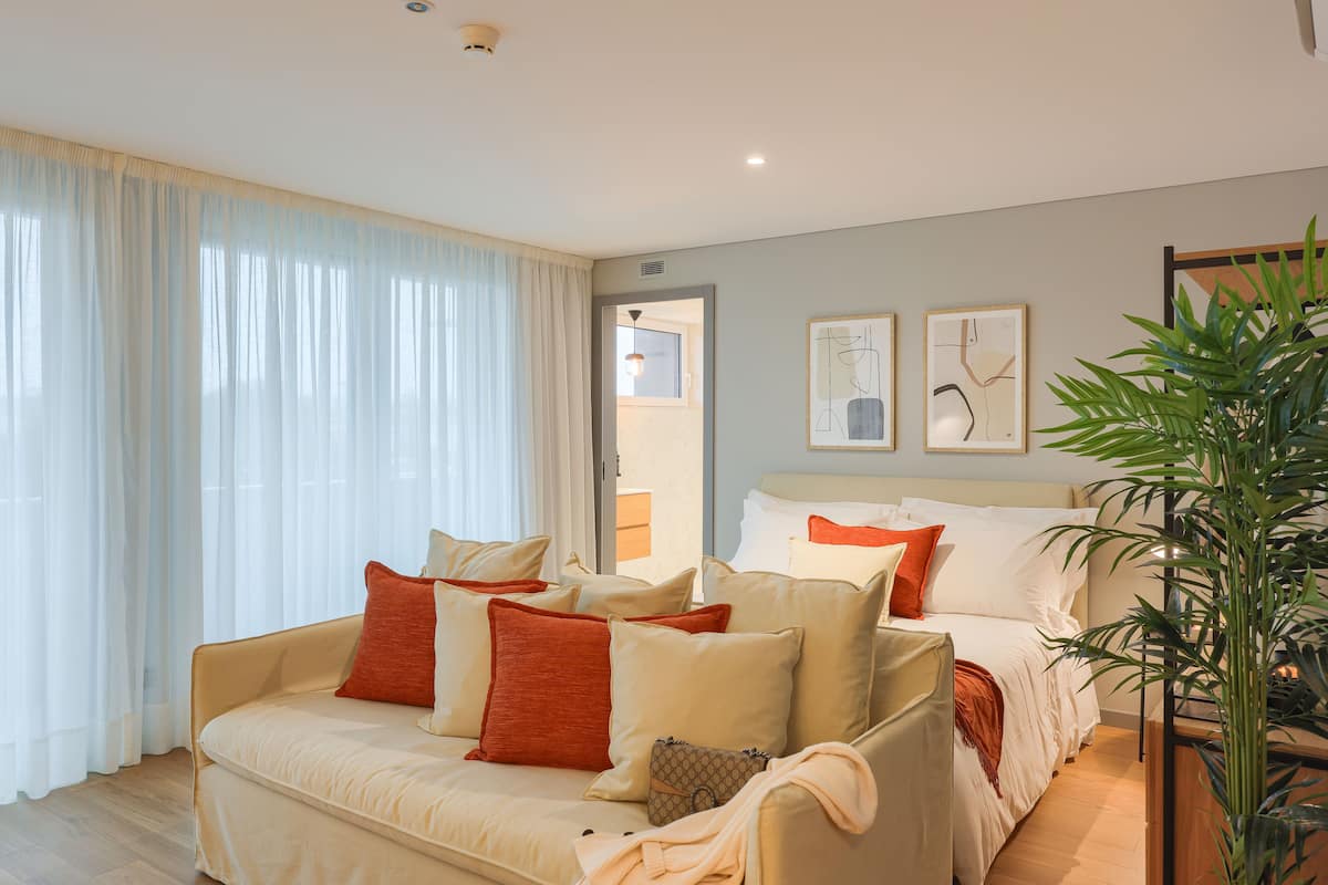 Una habitación de hotel moderna con un sofá beige, una cama con ropa de cama blanca y naranja, y una planta verde grande.