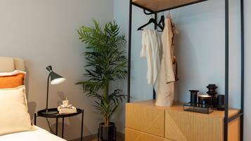 A neatly arranged hotel room with a bed, bedside table, lamp, potted plant, and a wooden dresser with a mirror and hanging clothes.