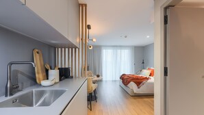 A modern kitchen with a sink, wooden cutting board, and utensils. A dining area with a table and chairs is visible. A bed with a red blanket is in the background. - Nôma Living Icon Apartments (Porto)