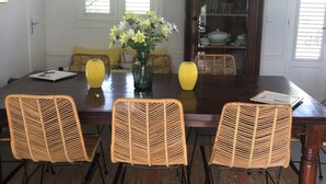 Uma área de refeições com uma mesa de madeira escura, cadeiras de vime, um vaso com flores e um lustre.
