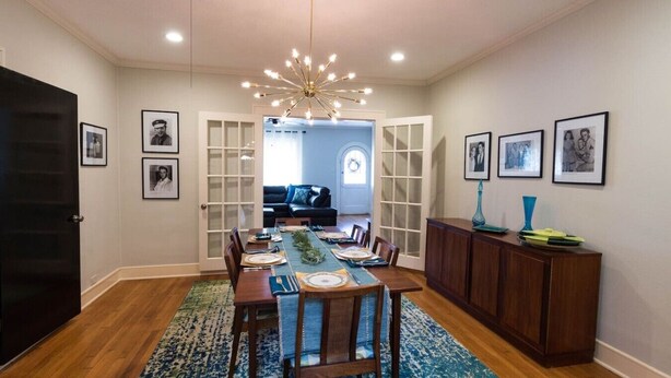 Comedor con mesa de madera dispuesta para seis comensales, una araña y vista a un salón a través de una puerta de vidrio.