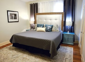 Une chambre à coucher avec un grand lit, une table de chevet, un tableau encadré au mur et une fenêtre avec des rideaux.