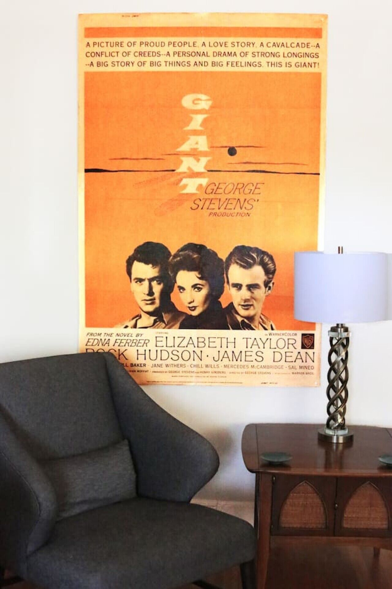 Un cartel de película en una pared, un sillón oscuro y una mesita de madera con una lámpara.