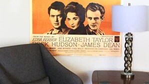 Un cartel de película en una pared, un sillón oscuro y una mesita de madera con una lámpara.