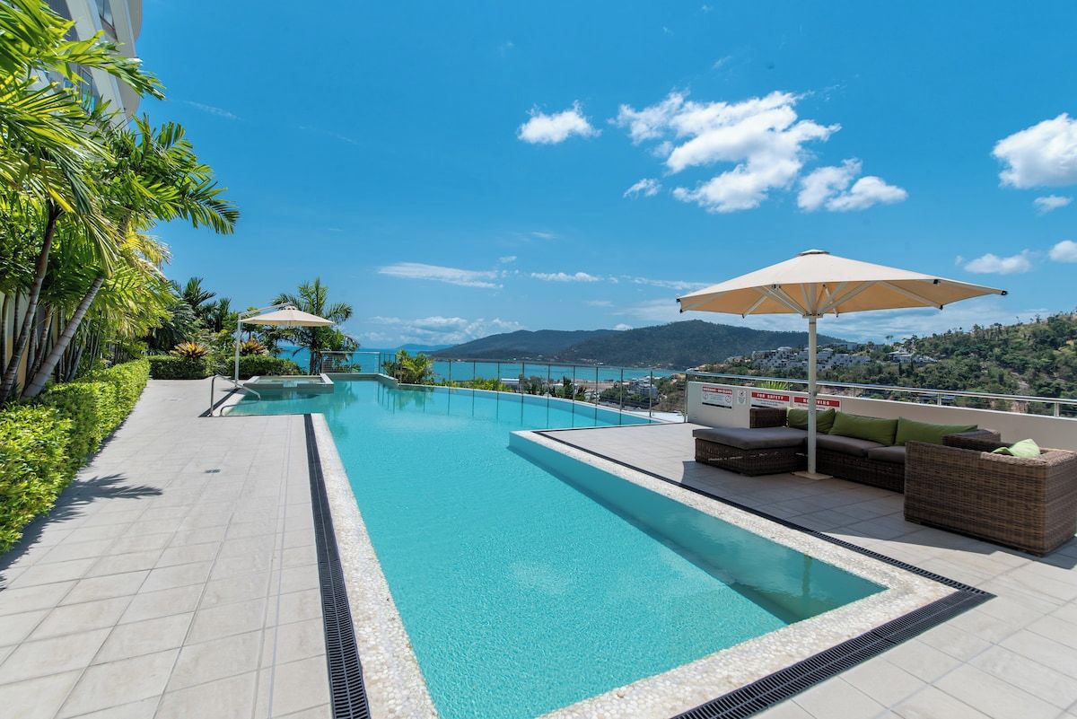 Une piscine avec vue sur la mer et les montagnes, entourée de chaises longues et de parasols.