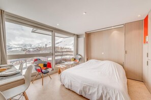 A modern hotel room with a large bed, a dining table, and chairs, a small table with a chair, and a view of a snowy mountain landscape.