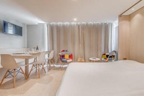 A modern hotel room with a large bed, a desk with chairs, a TV, and a window with curtains.
