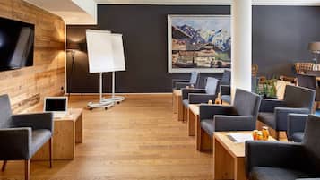 A modern hotel lobby with wooden flooring, a large painting of a mountainous landscape, and several seating areas with tables and chairs.