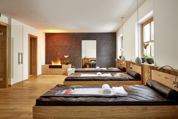 A modern spa room with wooden floors, multiple massage beds, a fireplace, and large windows. - Der grüne Baum Hotel (Ehrwald)