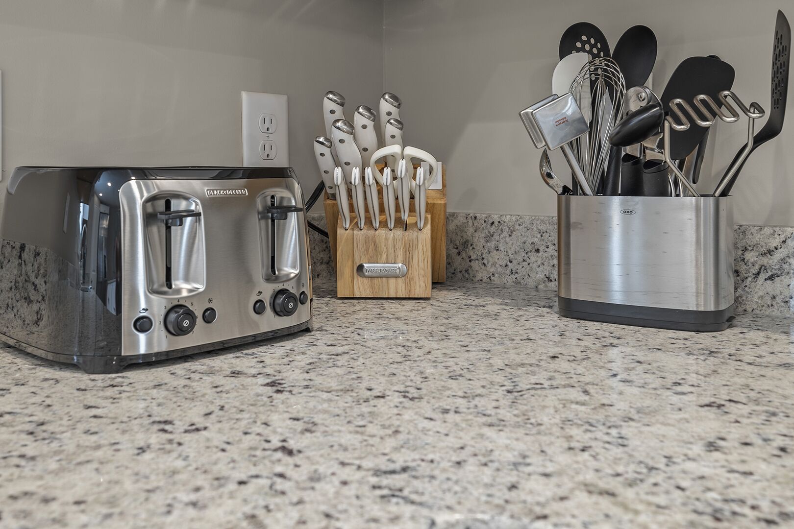 A kitchen with a stainless steel toaster, a knife block, and a utensil holder on a granite countertop.