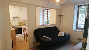 Un espace de vie compact avec un canapé noir, une petite table ronde et un coin cuisine visible à travers une porte ouverte.