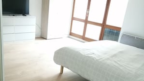 Une chambre à coucher avec un lit, un téléviseur posé sur un meuble blanc et une grande fenêtre donnant sur l’extérieur.