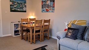 Ein Wohnzimmer mit Esstisch, Stühlen, einem Glastisch und einer Couch mit Kissen. An den Wänden hängen zwei Gemälde und eine kleine Ablagefläche mit Gegenständen.