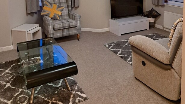 Ein Wohnzimmer mit Flachbildfernseher, einem Karoschen Sessel, einem Glastisch und einem beigefarbenen Sofa.