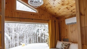A cozy wooden cabin room with a large window overlooking a snowy forest, a bed with a butterfly pillow, and a chandelier.