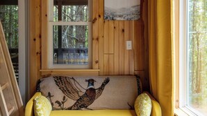 A wooden cabin interior with a yellow sofa, two patterned pillows, a framed picture on the wall, and a window with a view of trees.