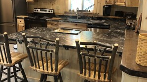 A kitchen with wooden cabinets, a stainless steel refrigerator, a stove, and a sink. There are three wooden chairs at the island. - Adirondack Home on Lake Snow (Indian Lake)