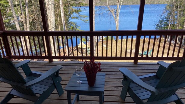A wooden deck with two Adirondack chairs and a small table with a glass of water and a vase of flowers. - Adirondack Home on Lake Snow (Indian Lake)
