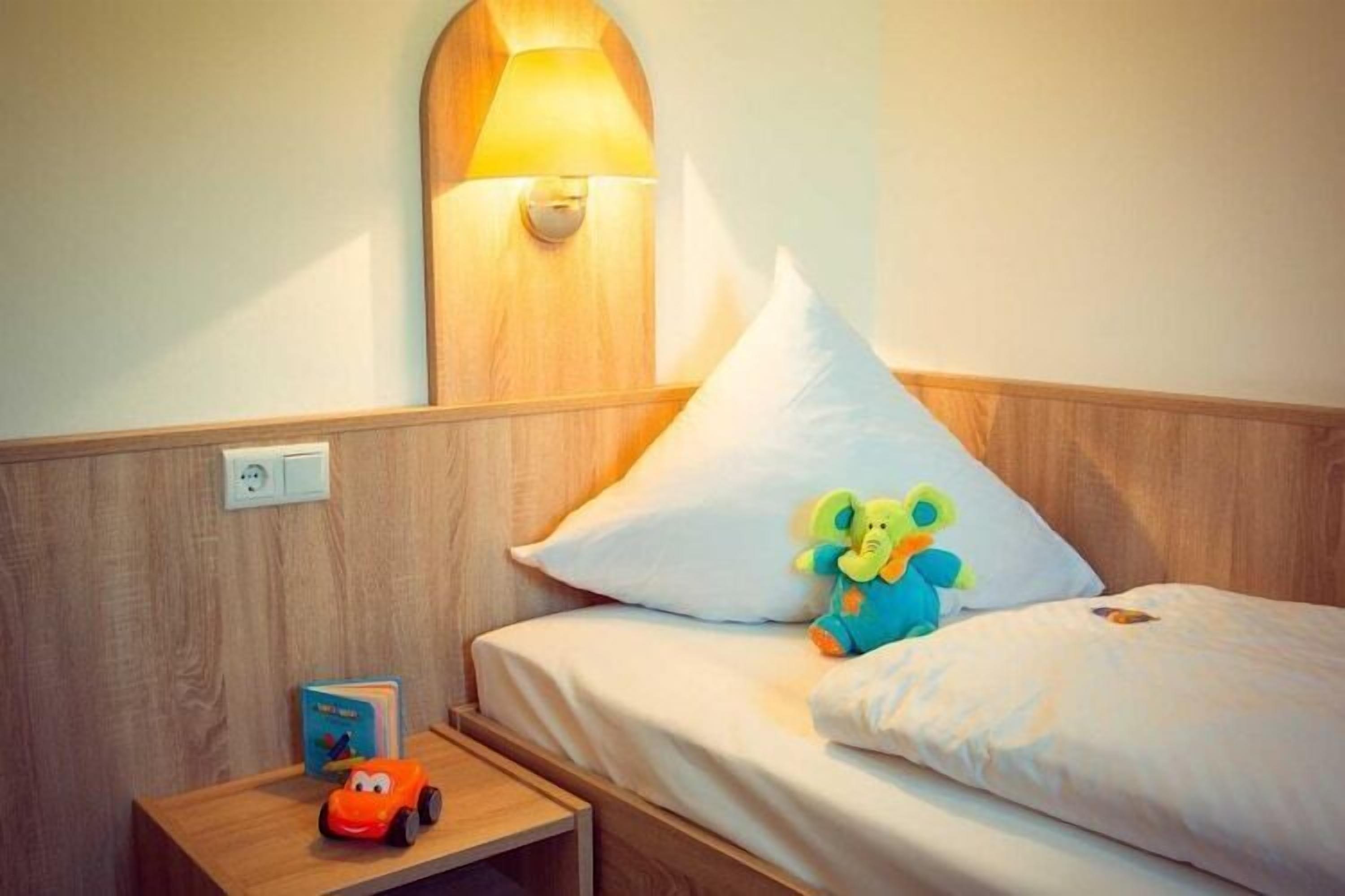A hotel room with a wooden headboard, a bedside lamp, a bed with white bedding, a plush toy elephant, and a toy car on the nightstand.