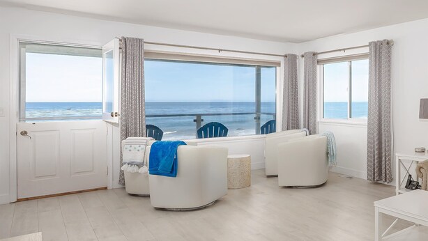 Une pièce dotée d’une grande fenêtre donnant sur la mer, d’un canapé blanc, d’une petite table ronde et d’une porte blanche.
