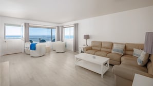Un salon moderne avec un canapé beige, des fauteuils blancs, une table basse blanche et une vue sur la mer grâce à de grandes fenêtres.