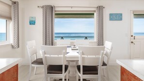 Un coin repas avec vue sur l’océan, des chaises blanches, une table et une fenêtre avec des rideaux.
