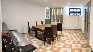 Un comedor con una mesa y sillas de madera, un sofá con funda estampada, piso de baldosas, una ventana con cortinas y una pantalla mostrando información.