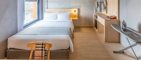 A modern hotel room with a large bed, a desk, and a chair. There is a window with blinds, a hanging light, and a small ironing board.