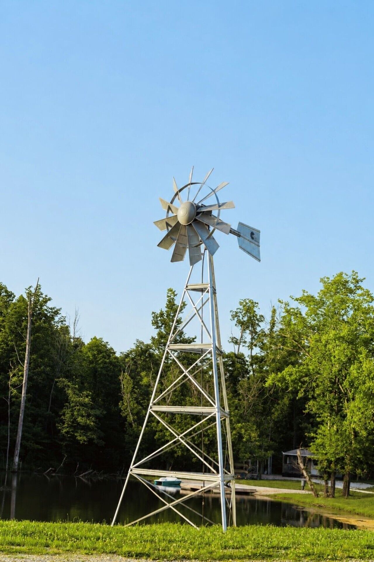 A windmill with a metal structure and blades, located near a body of water and surrounded by trees.