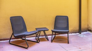 Dos sillas de mimbre y una pequeña mesa frente a una pared amarilla, con suelo de baldosas.