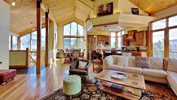 A spacious living area with a high ceiling, wooden beams, a large sofa, a coffee table, and a view of the kitchen and dining area.