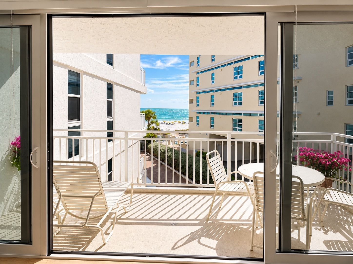A balcony with white metal furniture, a view of the beach, and buildings in the background.