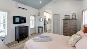A bedroom with a bed, dresser, TV, and a kitchenette in the background. - Charming Tiny Cottage, 5min to SAV, 15min to Beach by Lucky Savannah (Thunderbolt)