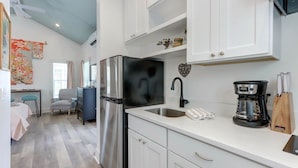 A modern kitchen with white cabinets, a stainless steel refrigerator, a coffee maker, and a heart-shaped decoration on the wall. - Charming Tiny Cottage, 5min to SAV, 15min to Beach by Lucky Savannah (Thunderbolt)