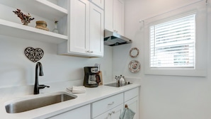A kitchen with white cabinets, a stainless steel sink, a coffee maker, and a kettle. There is a window with blinds and decorative plates on the wall. - Charming Tiny Cottage, 5min to SAV, 15min to Beach by Lucky Savannah (Thunderbolt)