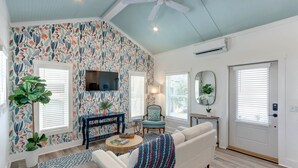 A living room with a patterned wallpaper, a ceiling fan, a flat-screen TV, a sofa, a coffee table, and a door with a window. - Marsh Hen Den - 5 Min to SAV / 15 to Beach by Lucky Savanah (Thunderbolt)