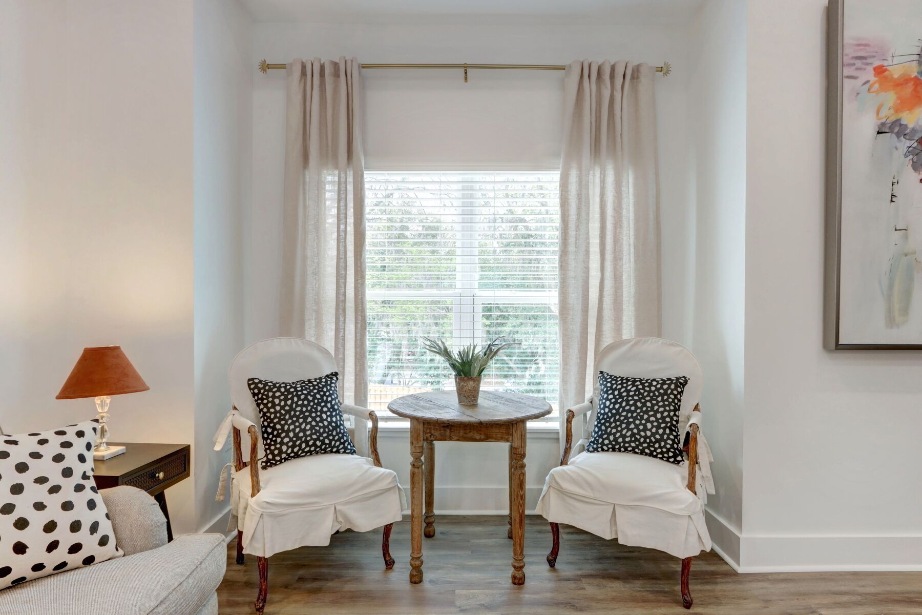 Ein Zimmer mit zwei weißen, gepolsterten Stühlen mit schwarz-weißen Polka-Dot-Kissen, einem kleinen runden Holztisch und einer Topfpflanze.