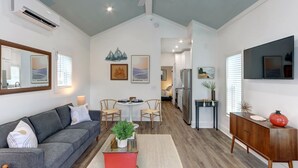 A spacious living area with a grey sofa, a red coffee table, and a dining table with chairs. There is a kitchen area with a refrigerator and a wooden cabinet.