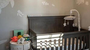 A baby crib with a dark headboard and a hanging mobile. A small table with toys and a green box is beside the crib.