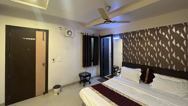 A hotel room with a bed, a ceiling fan, a small table, and a wall-mounted air conditioning unit. - Hotel Tushar Residency (Mathura)