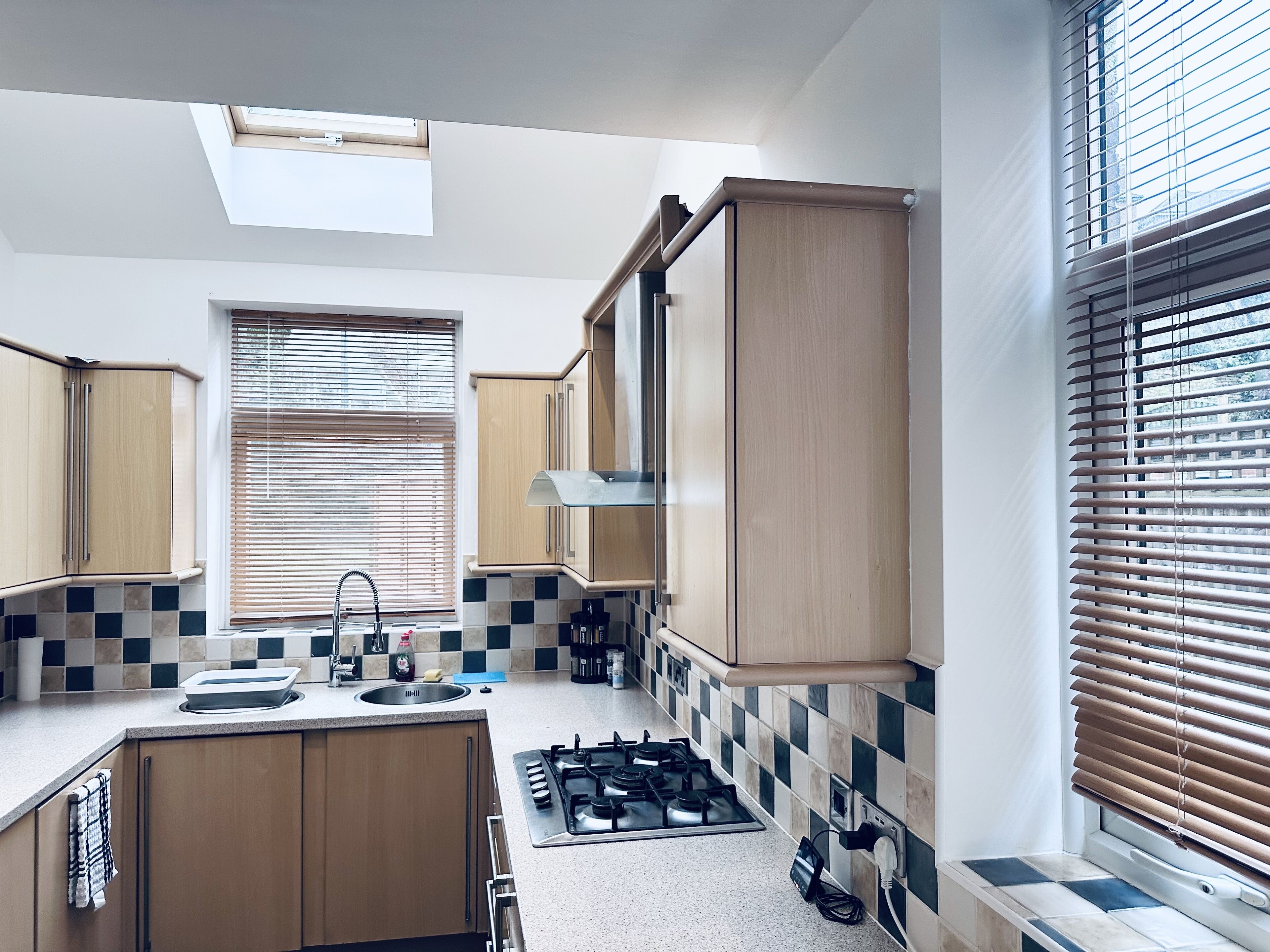 Une cuisine compacte équipée d’une cuisinière à gaz, de meubles en bois, d’une fenêtre avec des stores et d’un puits de lumière.
