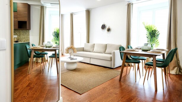 Una sala de estar moderna con sofá, mesa de comedor y sillas. Se ve un área de cocina a través de una puerta abierta.