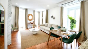 Una sala moderna con un sofá, una mesa de comedor y sillas. Hay un espejo grande en la pared, una planta y una ventana con cortinas.